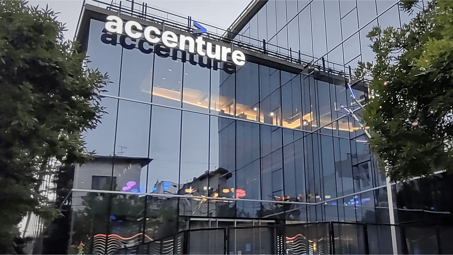 Accenture logo on the glass facade of a modern office building with reflections of nearby buildings and trees.