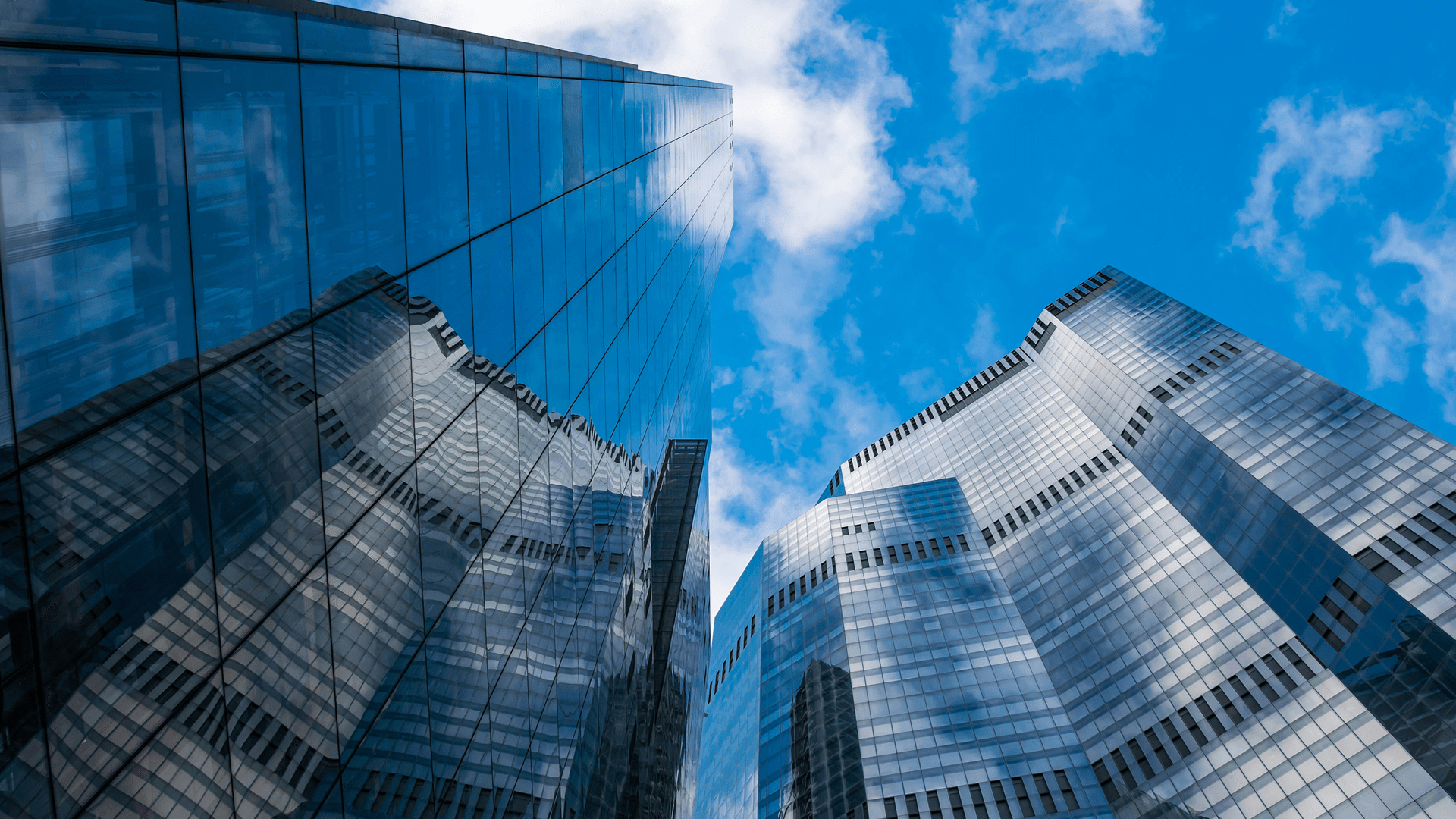 Upward view of tall glass skyscrapers reflecting blue sky and clouds.